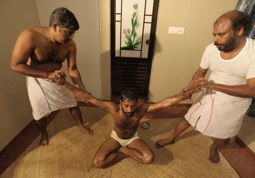 Two Marma therapists stretching both arms of a seated patient — carpal tunnel syndrome treatment at Agasthya, Kerala